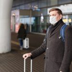 Young European man in gray coat, protective disposable medical mask in airport. Afraid of dangerous N-CoV 2019 influenza coronavirus mutated and spreading in China. Blue backpack, suitcase on wheels